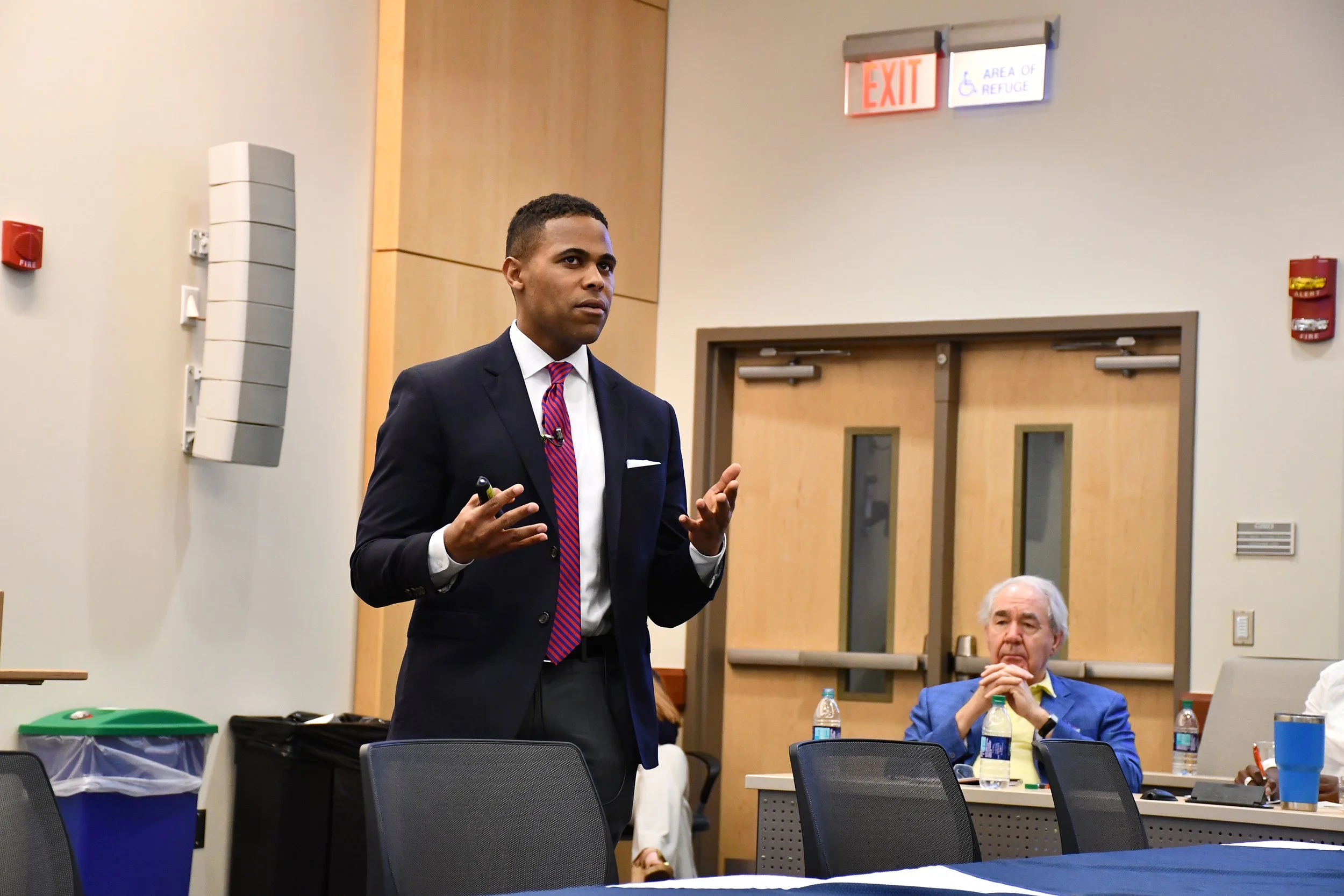 A man in a dark suit with a red and blue striped tie giving a presentation in a conference room, with a woman sitting at a desk listening in the background.