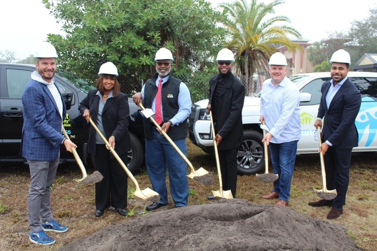 Group of six diverse professionals in suits and hard hats at a groundbreaking ceremony, holding shovels with dirt, smiling, with vehicles and trees in the background.