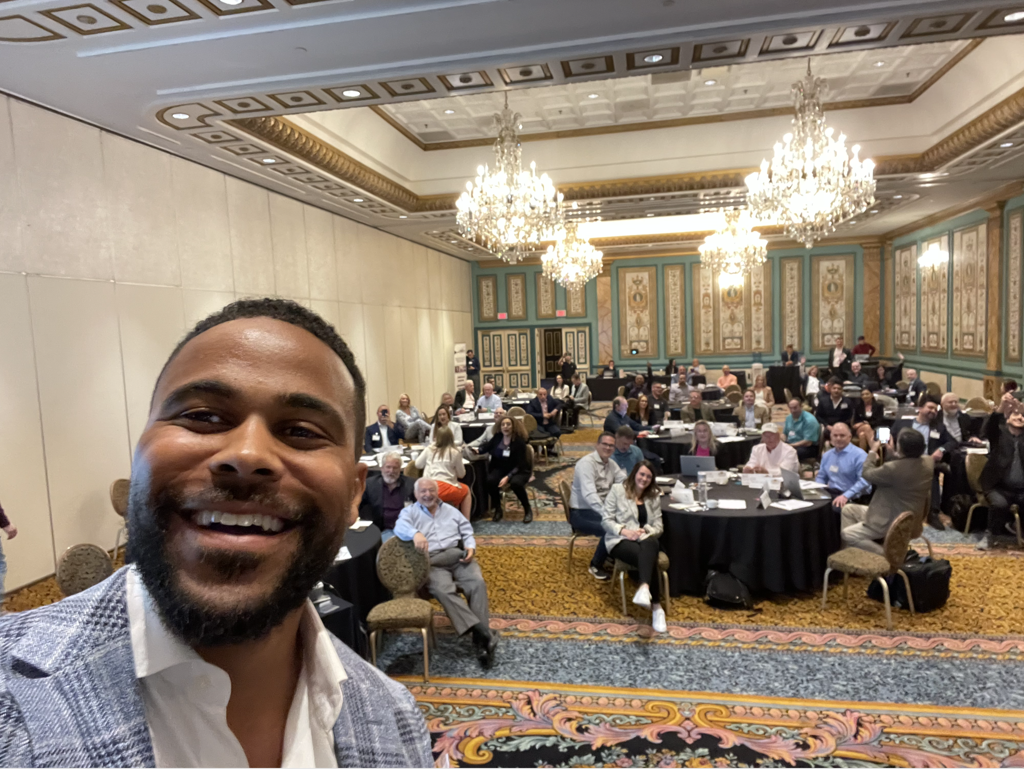 A man taking a selfie in front of a large conference room filled with people sitting at round tables under ornate chandeliers and decorated walls.