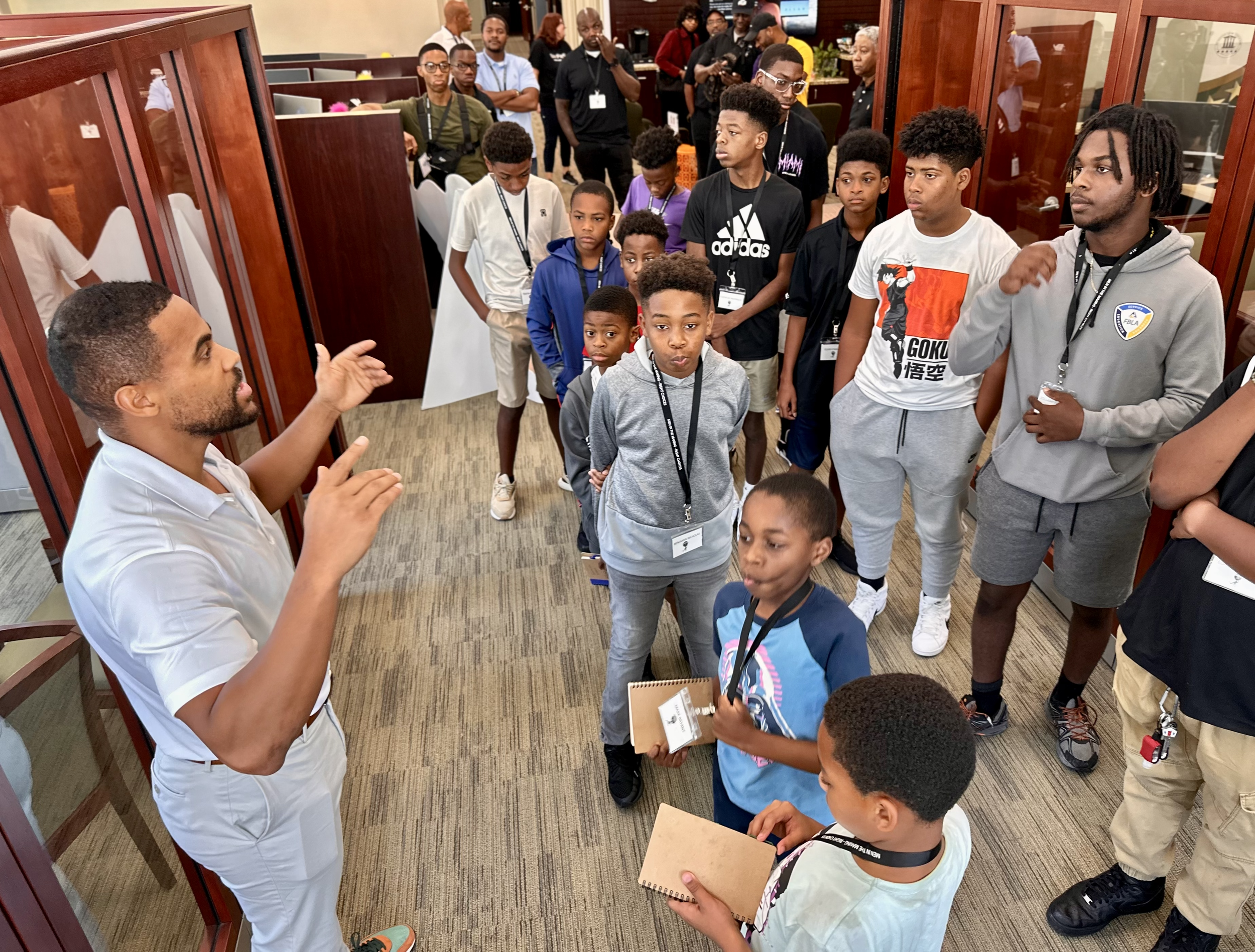 A man is giving a presentation to a group of children and young adults in an indoor setting, possibly a conference or educational event. The group is listening attentively.