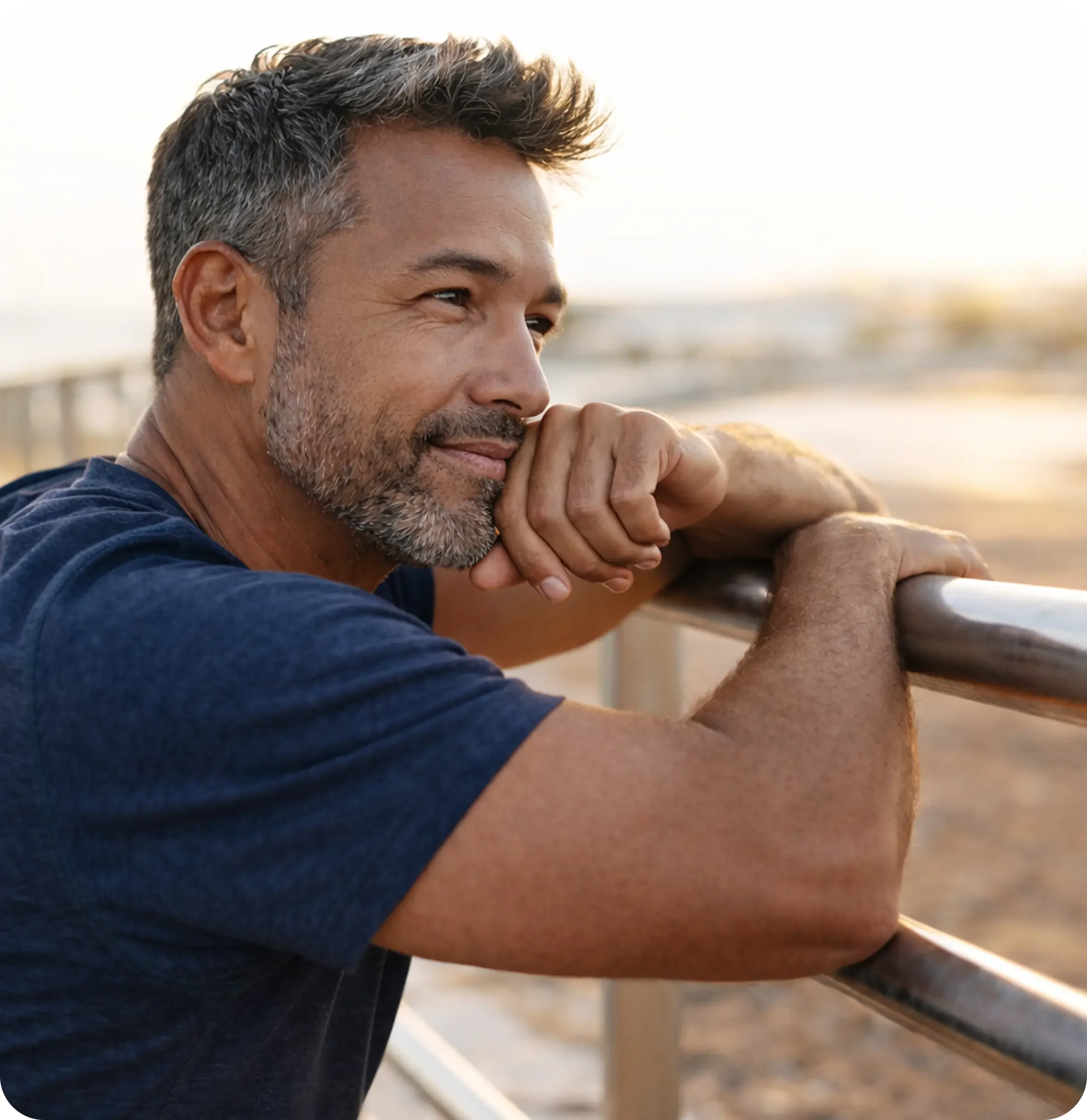 A middle-aged man with gray hair and a beard leaning on a metal railing at the beach during sunset, looking thoughtful.