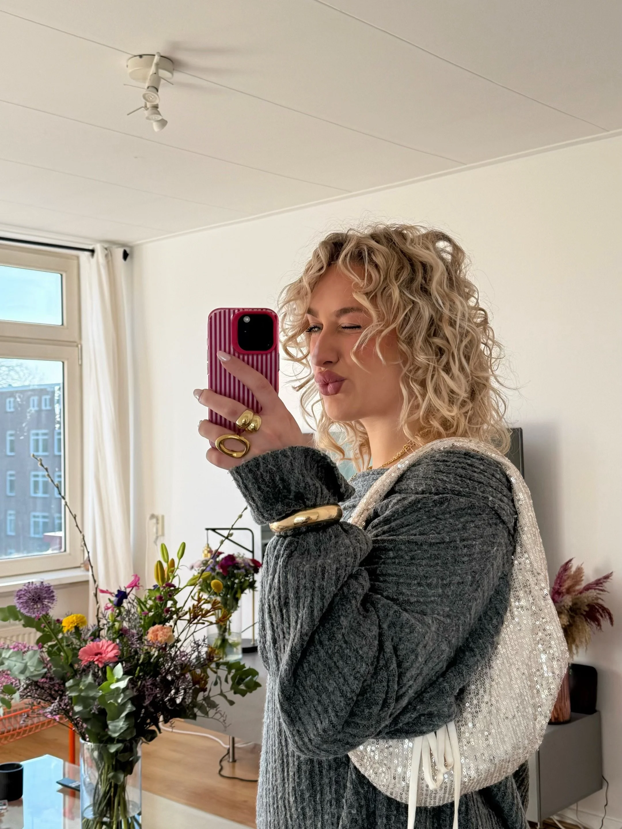 A woman with curly blonde hair taking a mirror selfie in a living room, holding her phone with a pink striped case. She is wearing gold jewelry and a dark gray sweater, with a silver sequin bag over her shoulder. There is a bouquet of colorful flowers on a table behind her, and a window with white curtains in the background.