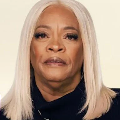 Close-up of an older woman with long, straight platinum blonde hair, wearing a dark top, against a light background.