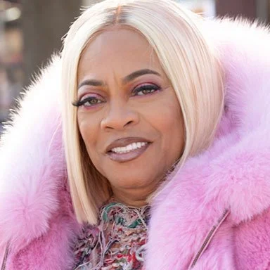 Close-up of a woman with blonde hair, wearing a pink faux fur coat with a hood, smiling slightly.