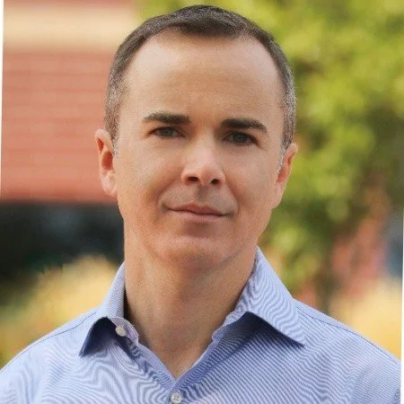 Portrait of a man in a blue dress shirt standing outdoors with a blurred background of greenery and a building.