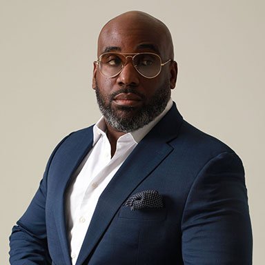 A man with glasses, wearing a navy suit and white shirt, standing against a plain background.