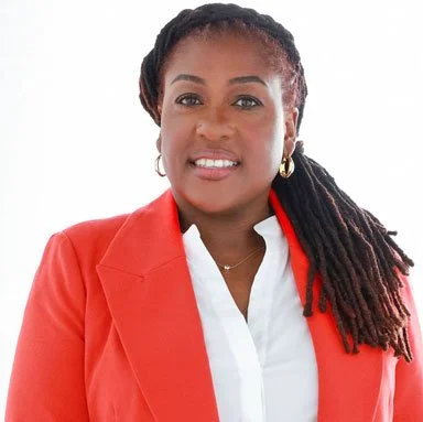 A woman with long dreadlocks, wearing a red blazer over a white shirt, smiling at the camera.