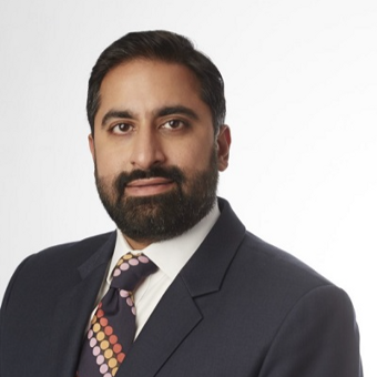 Professional headshot of a man with dark hair and beard, wearing a dark suit, white shirt, and patterned tie against a white background.