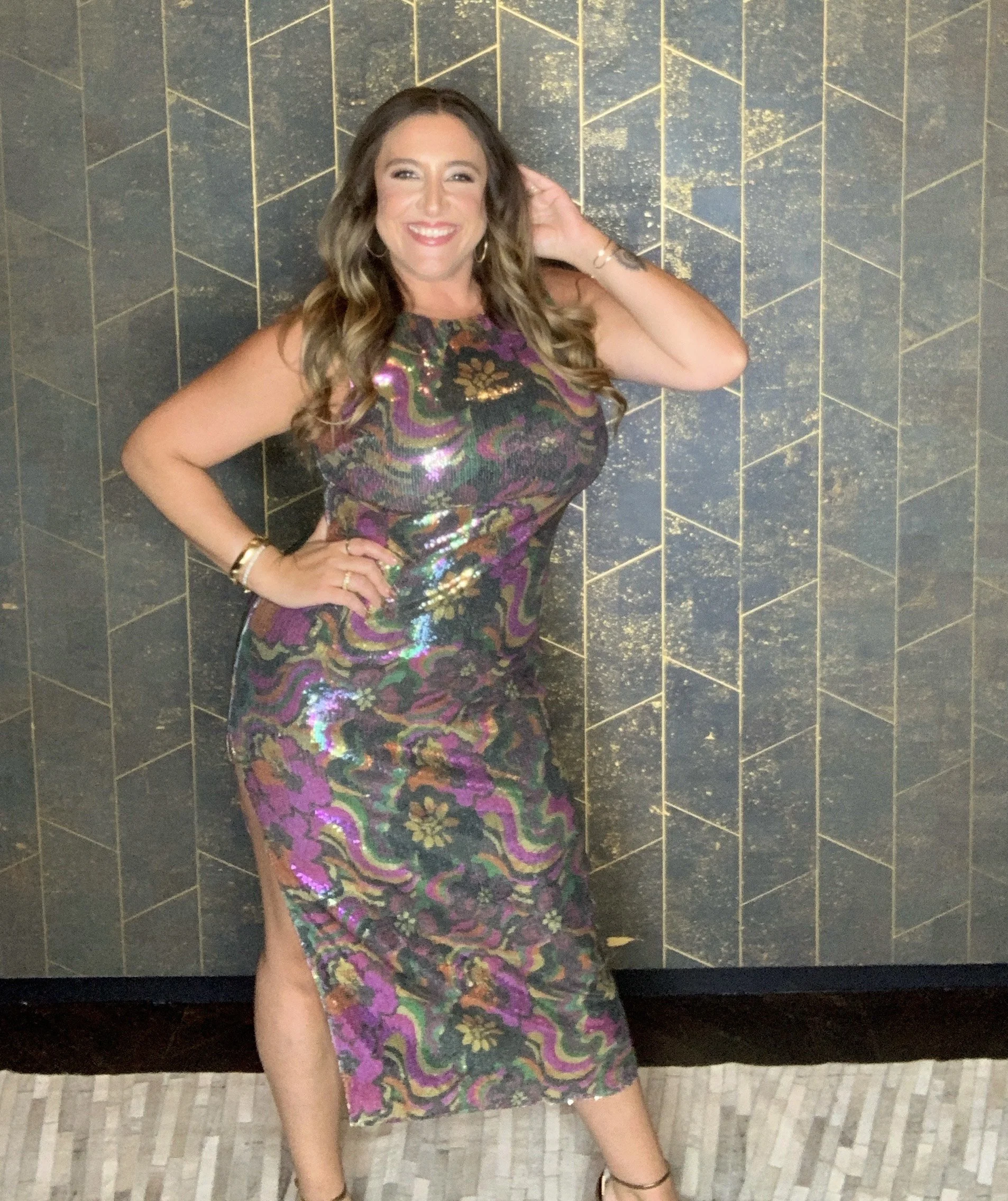 A woman in a colorful, shiny, patterned dress with a slit, smiling with one hand on her hip and the other touching her hair, standing in front of a decorative black and gold geometric wall.