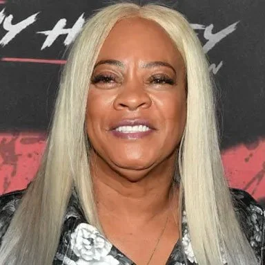 A woman with long, straight, platinum blonde hair smiling in front of a black and red background.