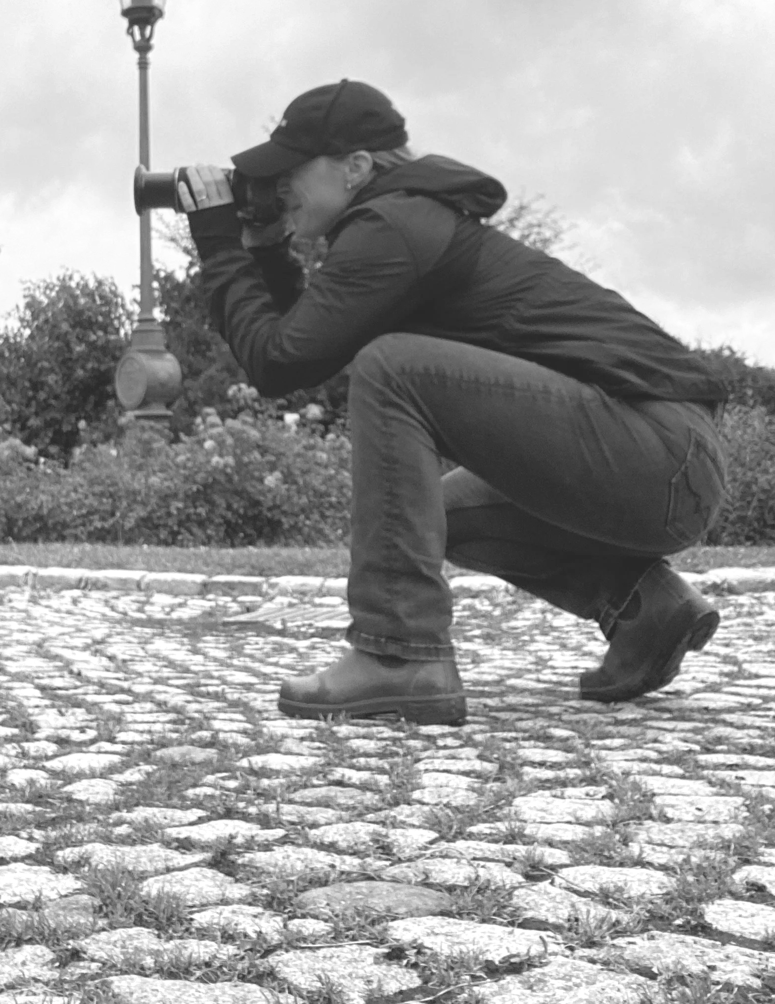 A person in outdoor clothing crouches on a cobblestone path, looking through binoculars.