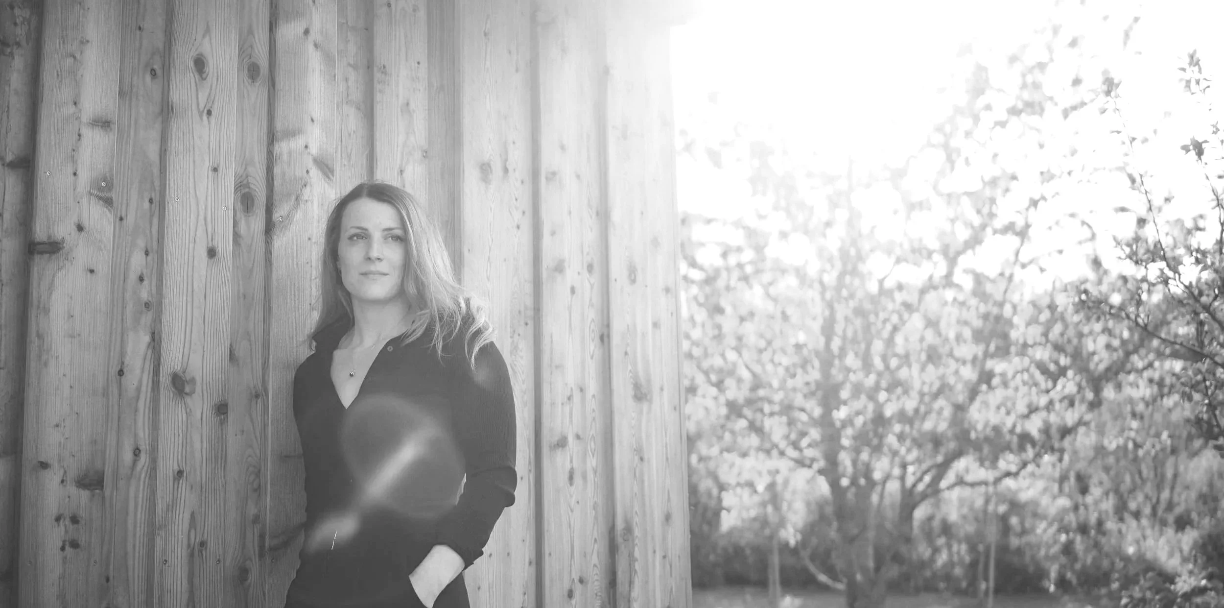 A woman with long hair standing against a wooden wall outdoors, with trees and branches in the background.