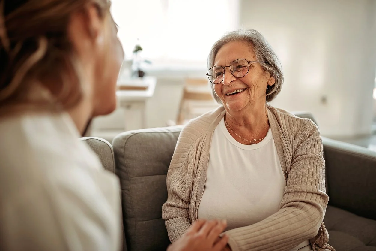 An elderly woman with glasses smiling and holding hands with another woman seated on a sofa in a bright living room.