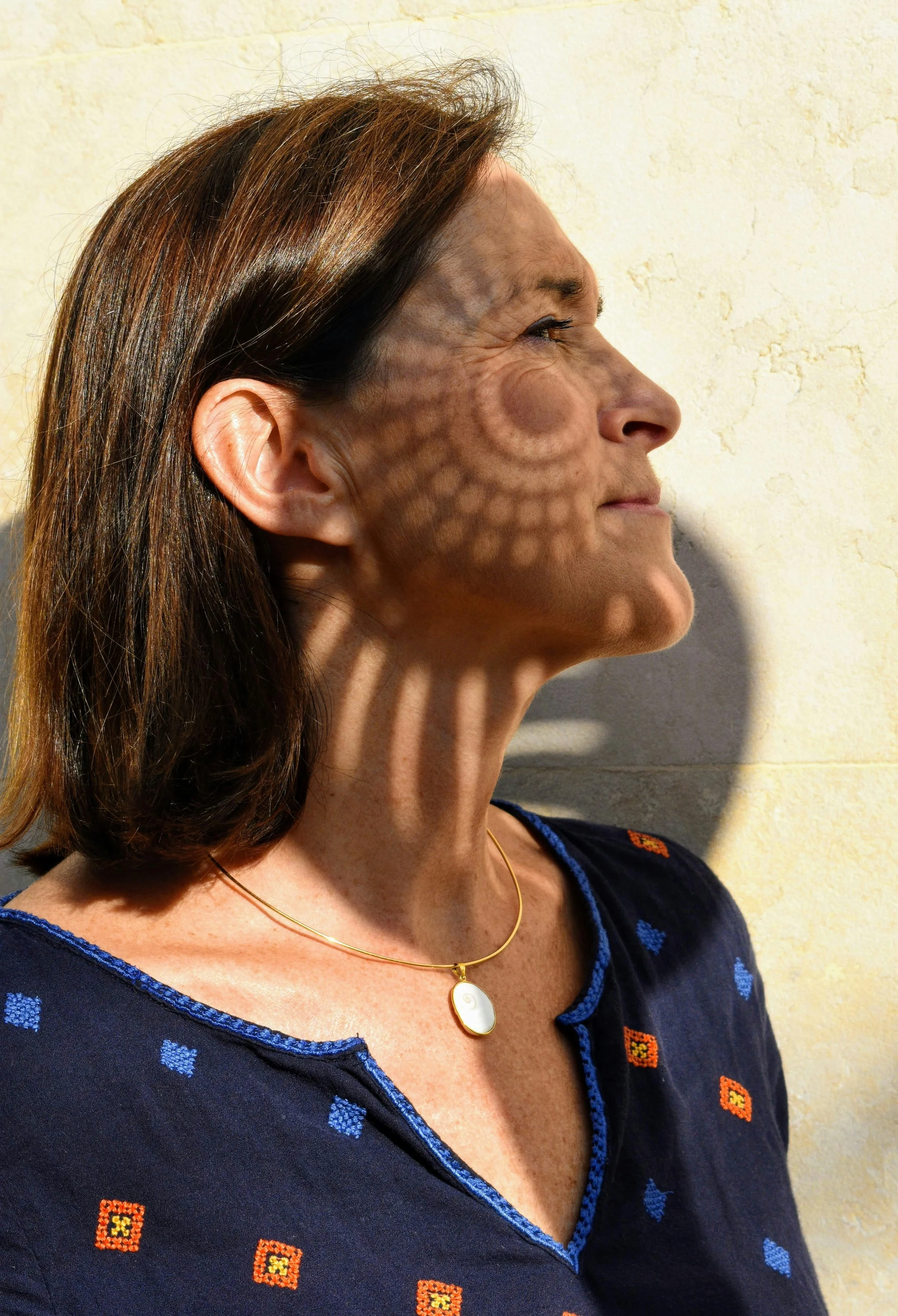A woman with brown hair is standing sideways against a wall, with sunlight casting a circular and striped shadow from a patterned object onto her face and neck.