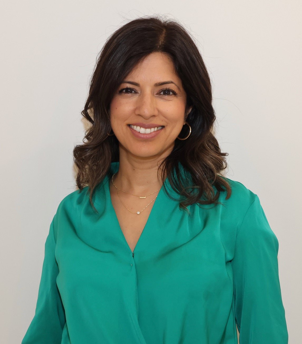A woman with shoulder-length dark hair, smiling, wearing a teal blouse and gold jewelry, standing against a plain white background.