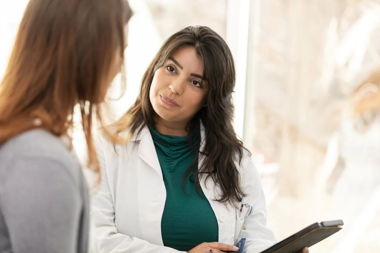 A female healthcare professional with dark hair and a green shirt, wearing a white lab coat, talking to a patient with reddish-brown hair in a gray sweater, holding a tablet device, near a window.