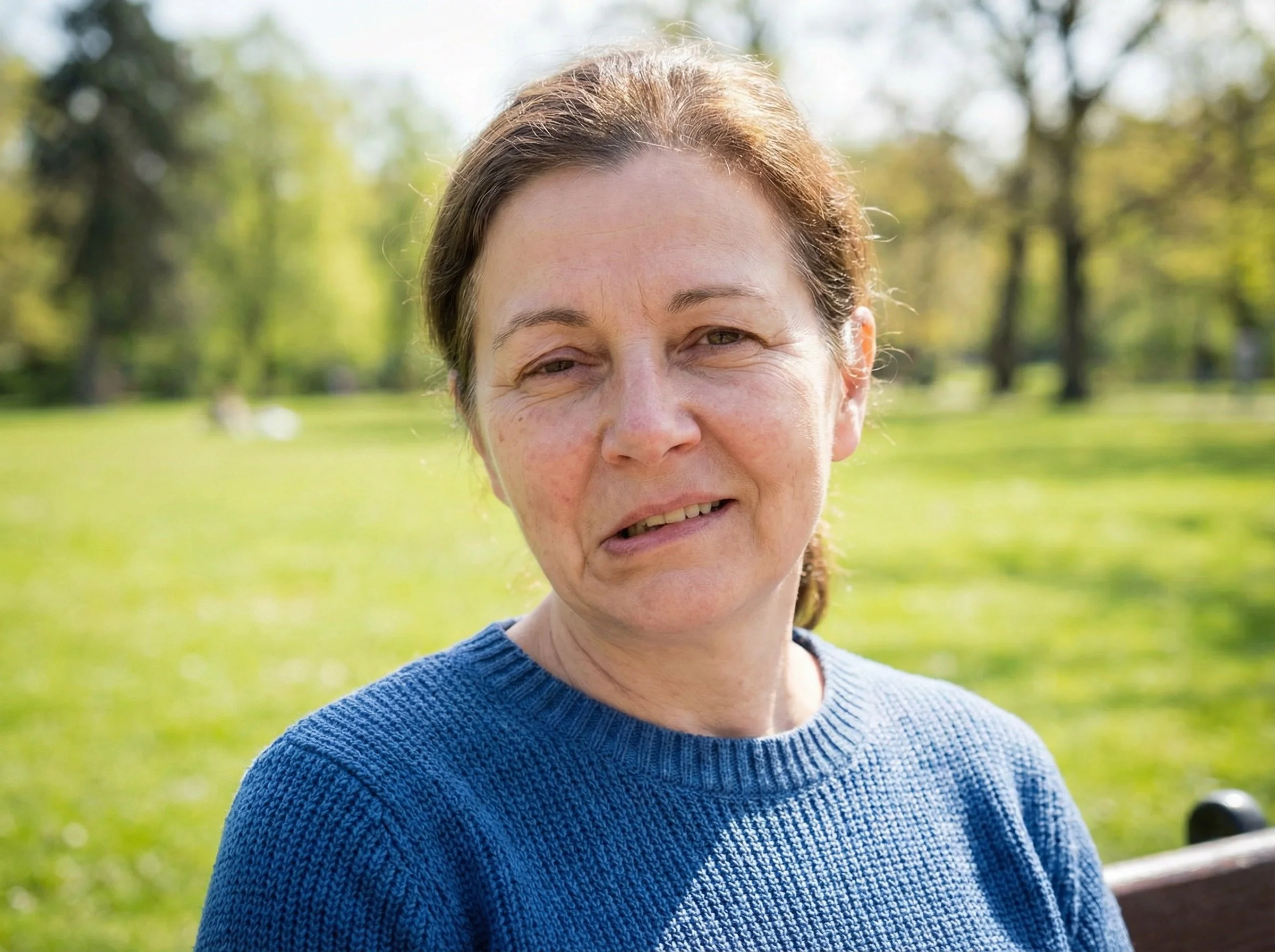 A woman with brown hair and a blue sweater sitting on a bench in a park with green grass and trees in the background, smiling at the camera.
