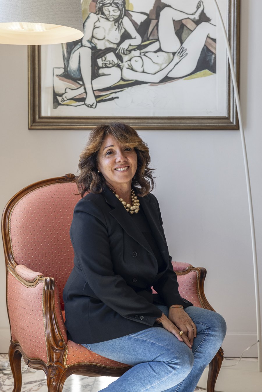 Una donna seduta su una poltrona rosa con motivo a pois, che sorride. Dietro di lei un dipinto di uno stile astratto con figure umane nude.