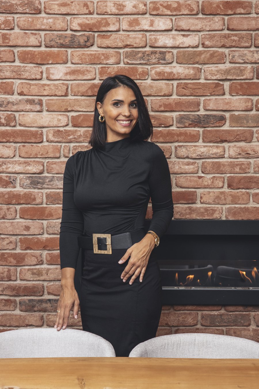 Una donna sorridente con capelli neri, vestita in abbigliamento nero, indossa orecchini dorati e un orologio, si trova accanto a un camino a parete di mattoni rossi.