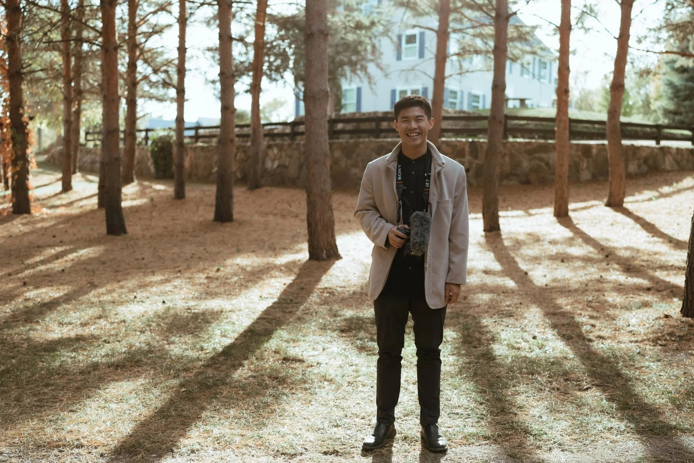 A young man smiling and holding a video camera in a wooded outdoor area with trees and a house in the background.