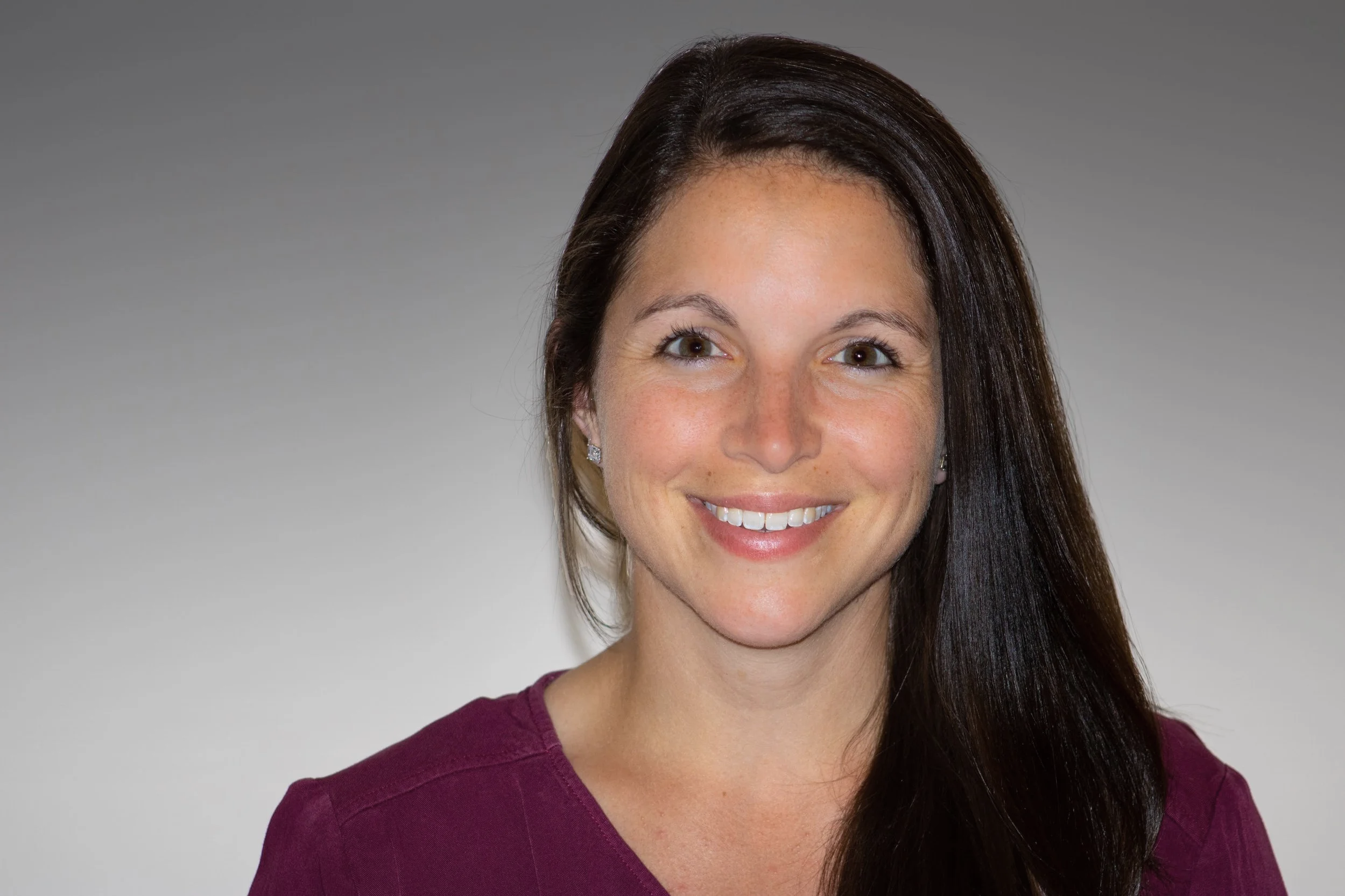 A woman with long dark brown hair, smiling, wearing a burgundy top, standing against a plain gray background.
