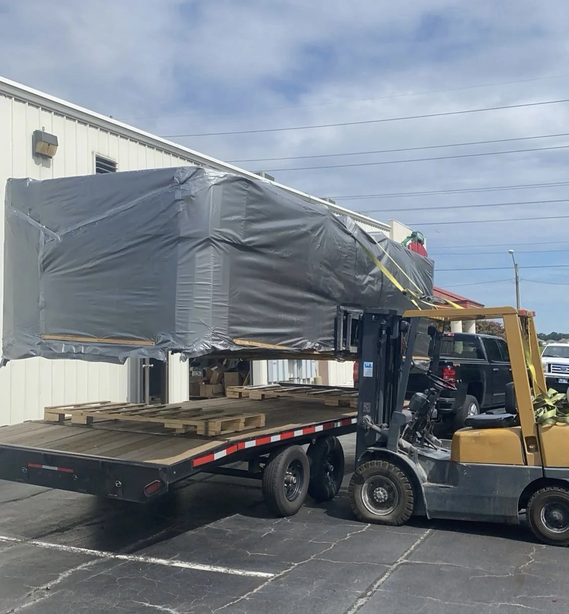 A forklift lifting a large, wrapped object onto a flatbed trailer outside a building.