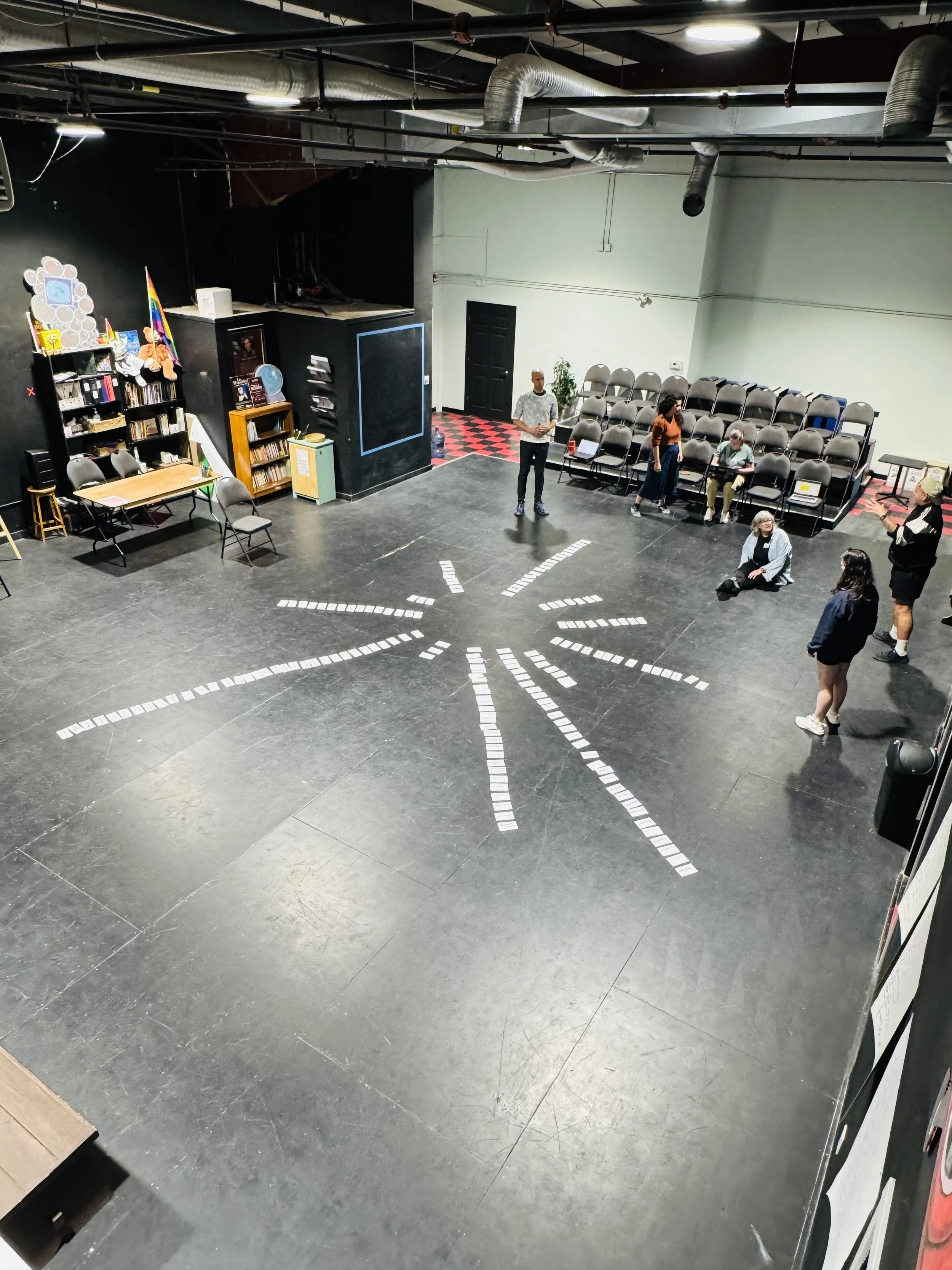 Theater stage with white rectangular cards arranged in a starburst pattern on the black floor, several people standing and sitting, stage area with bookshelves, chairs, and colorful decorations in the background.
