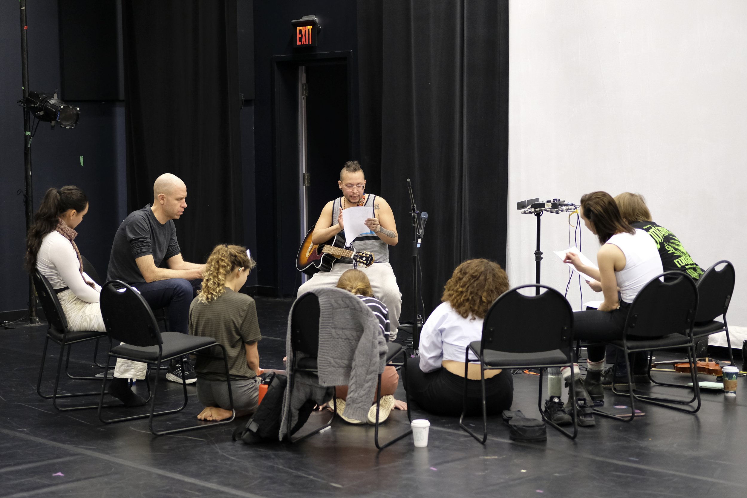 Group of people sitting on chairs and the floor in a rehearsal space with a musician holding a guitar and reading from a sheet of paper, surrounded by audio equipment and scripts.