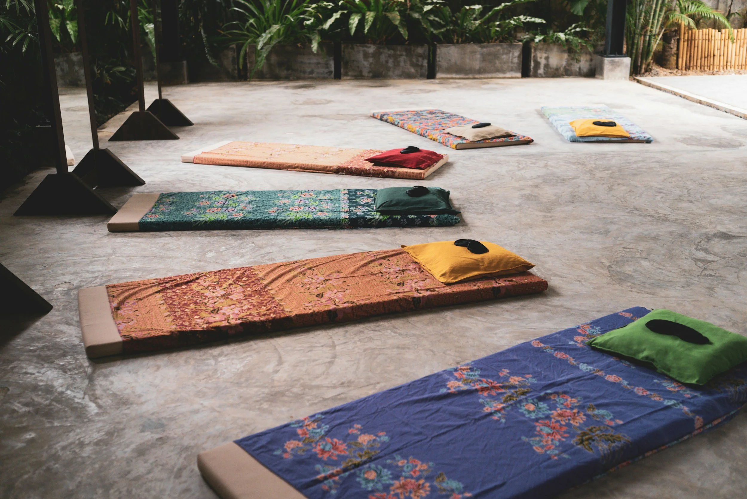 Multiple colorful yoga mats with pillows laid out on a concrete floor in an outdoor setting surrounded by plants.