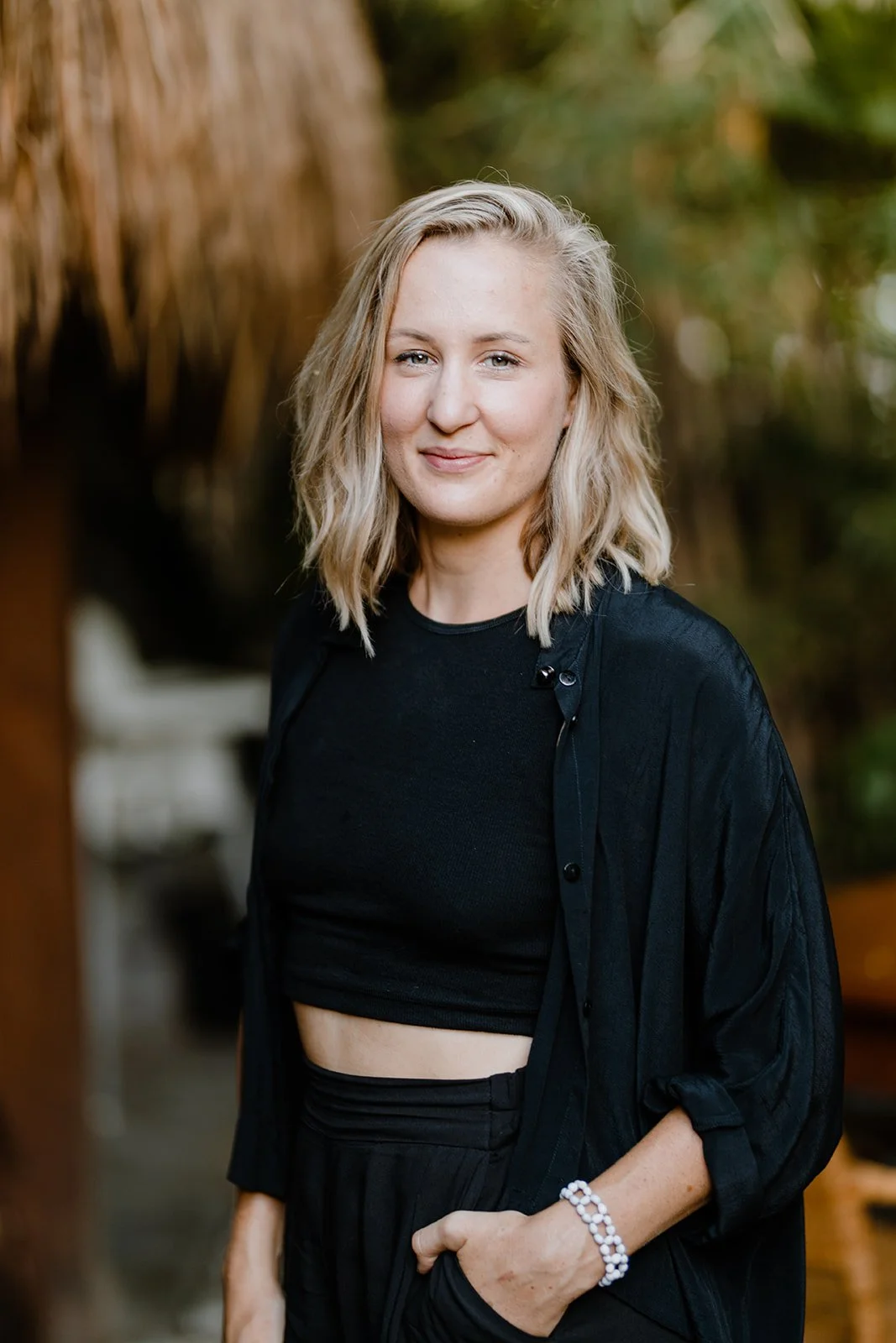 Jenny, with blonde, wavy hair, wearing a black crop top and black jacket, standing outdoors with blurred green and brown background.