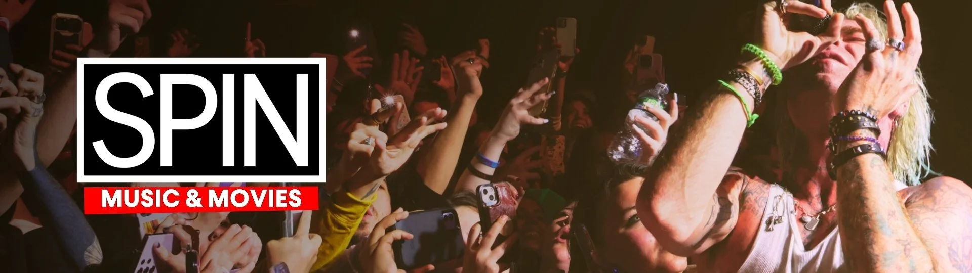 A singer with long blonde hair, tattoos, and multiple bracelets and rings is performing on stage, with fans taking photos and videos of her in the background. The text overlay reads 'SPIN MUSIC & MOVIES'.