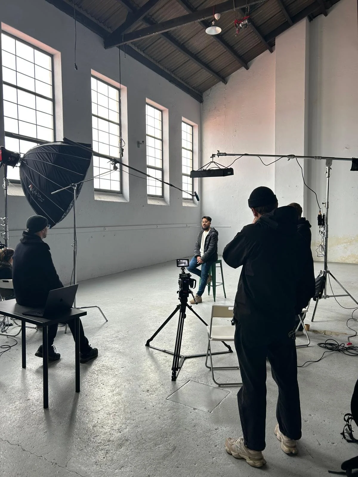 A man in a gray jacket sitting on a green stool in a photo shoot setup, surrounded by lighting and camera equipment, in a spacious studio with large windows and white walls.