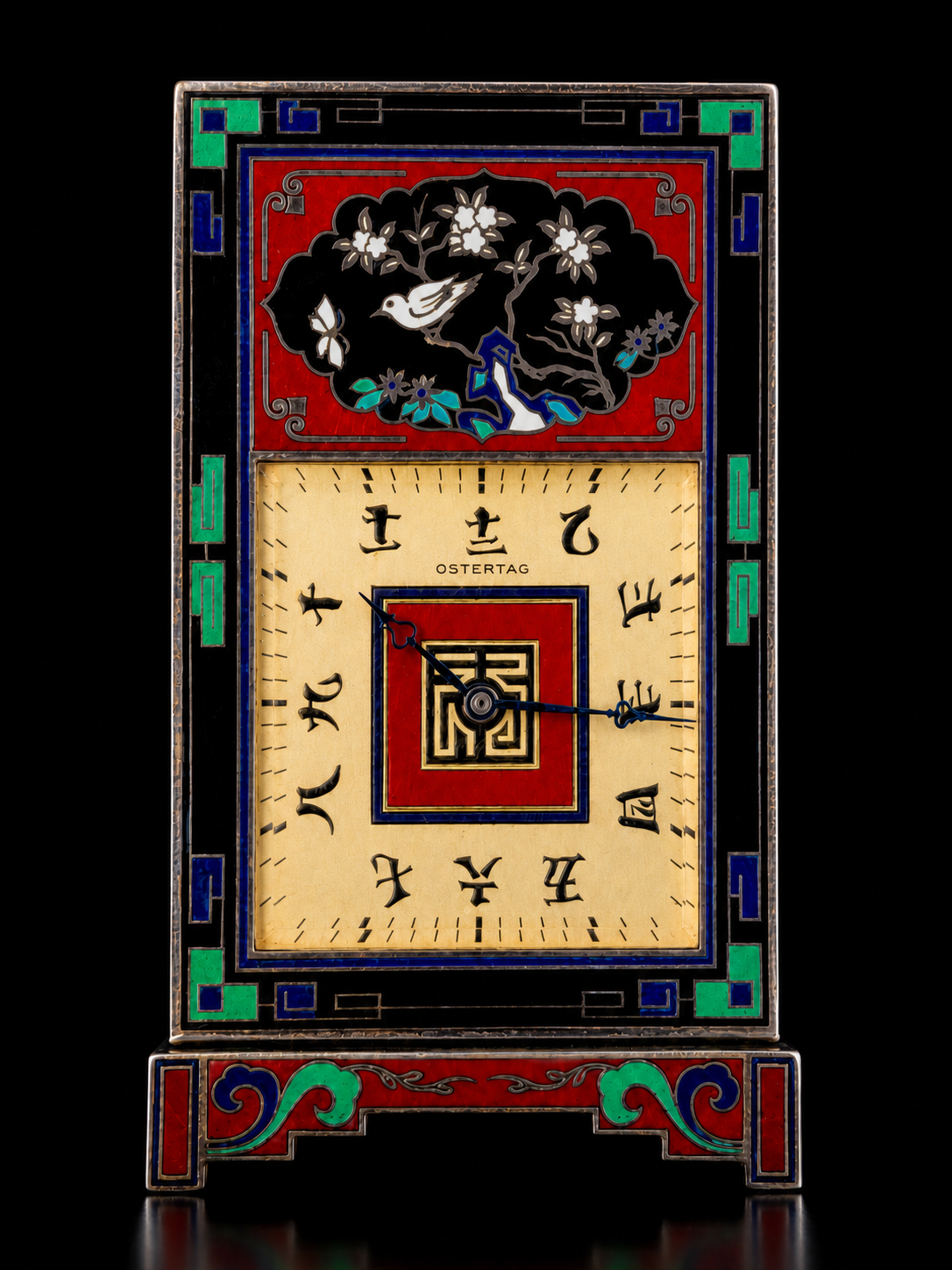 Une horloge en verre coloré avec des motifs floraux et un oiseau en haut. Le cadran est oval avec des chiffres stylisés pour l'heure. Les couleurs dominantes sont le rouge, le noir, le beige, le vert, le bleu et l'or.