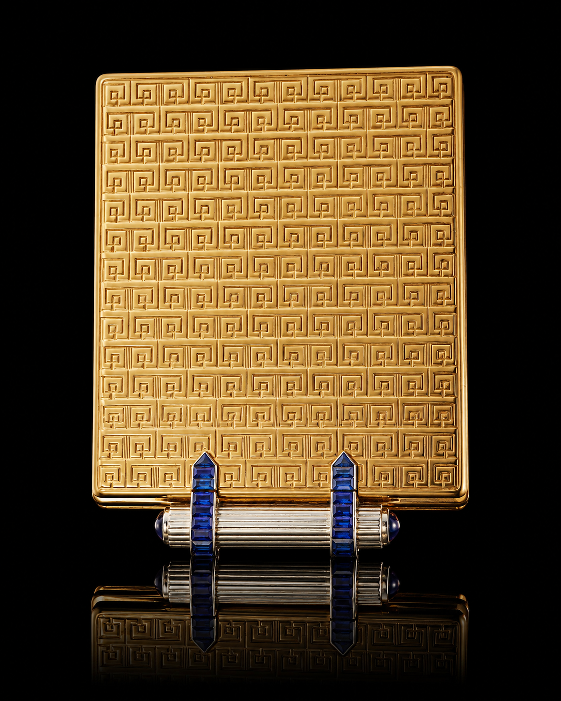 Un étui à cigarettes doré avec un motif géométrique, posé sur un fond noir. Il est soutenu par deux supports décorés de pierres bleues.