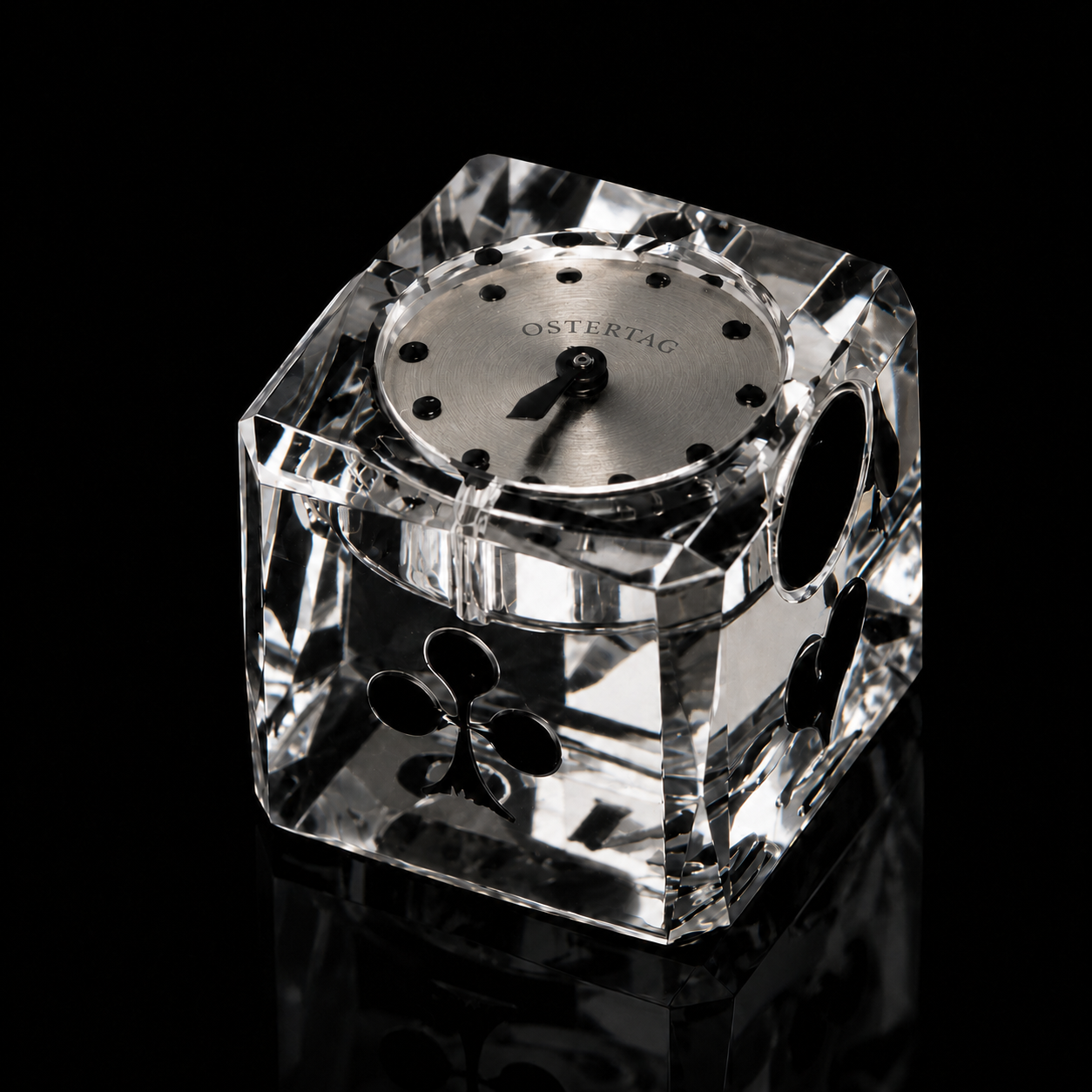 Montre avec cadran argenté portant l'inscription OSTERTAG, placée dans une boîte en cristal en forme de cube, décorée de trèfles noirs sur chaque face, sur un fond noir.