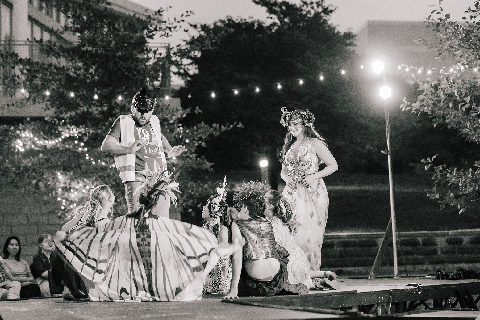 Performers in costumes standing and sitting on a stage during an outdoor nighttime performance, with audience members seated in the background and string lights overhead.