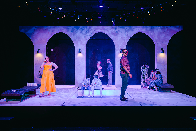 Actors on stage during a theatrical performance, with a backdrop of arches and string lights, some seated and others standing in various poses and costumes.