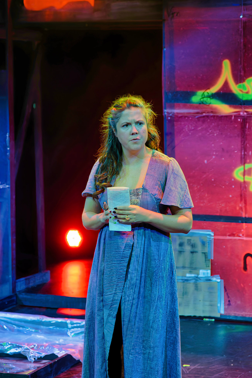 A woman with long curly hair holding a piece of paper, standing on a stage with colorful lighting and abstract background.