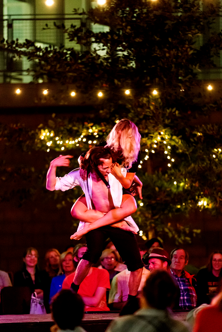 Performers dancing on stage at night, with audience watching, outdoors with string lights and trees in the background.
