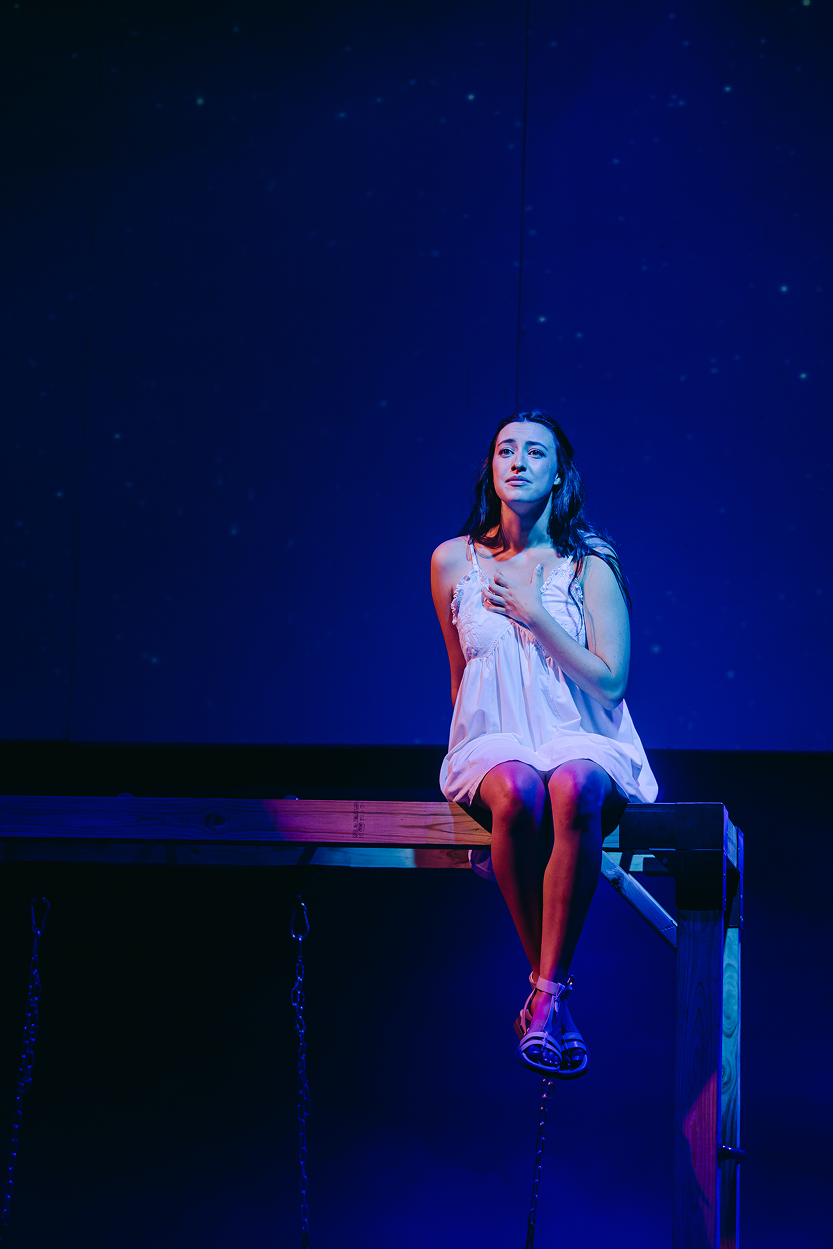 A woman sitting on the edge of a wooden platform or stage, dressed in a white nightgown, with a solemn or emotional expression, illuminated by blue stage lighting, with a starry sky projected on the background