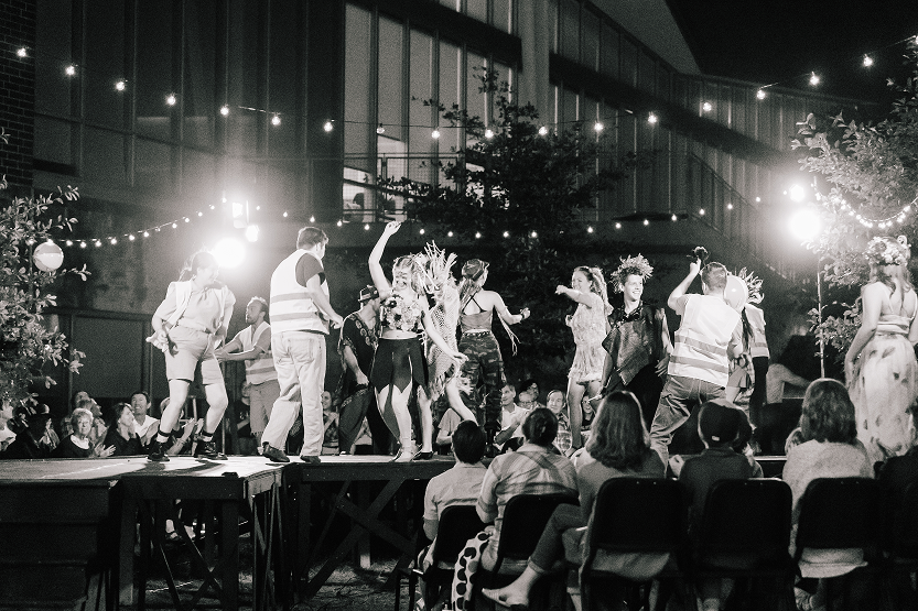People dancing on an outdoor stage at night during a lively celebration, with string lights overhead and an audience seated in front.