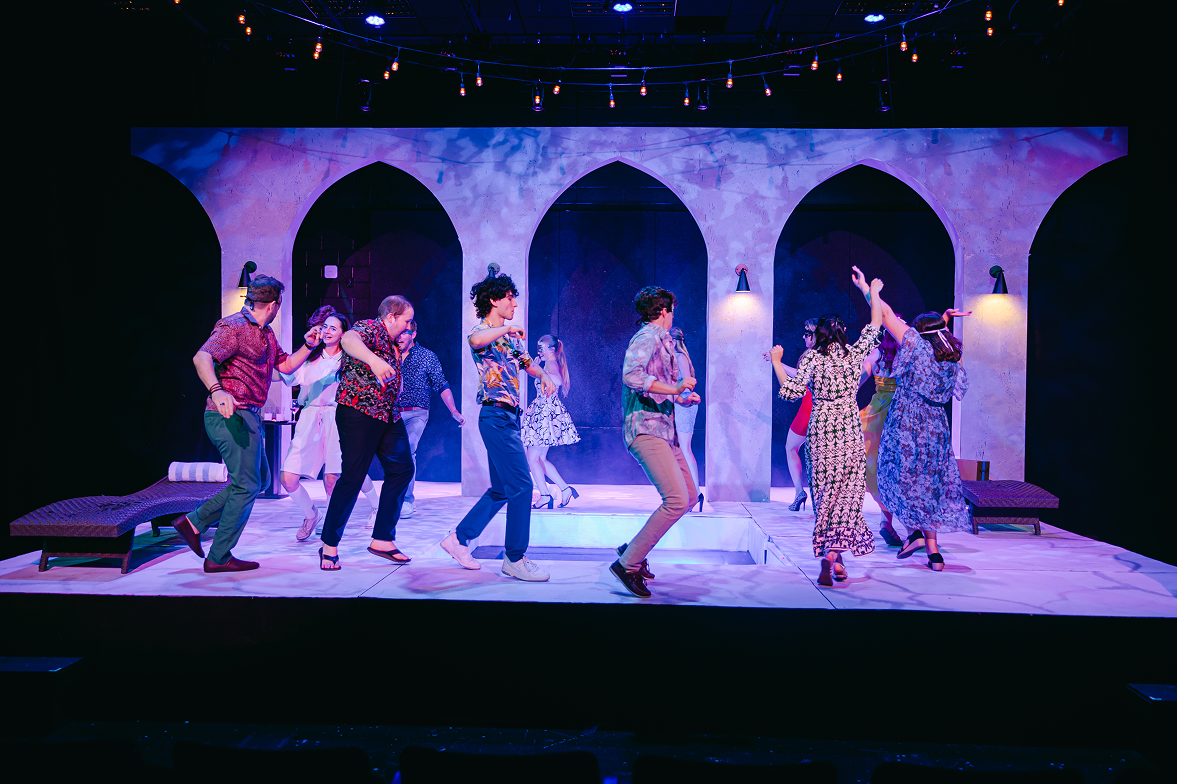 The image shows a stage play with multiple actors dancing and engaging in lively activities. The stage has a Mediterranean-style backdrop with arches and wall lamps, and there are lounge chairs on either side. The scene is lit with colorful lighting, creating a vibrant atmosphere.