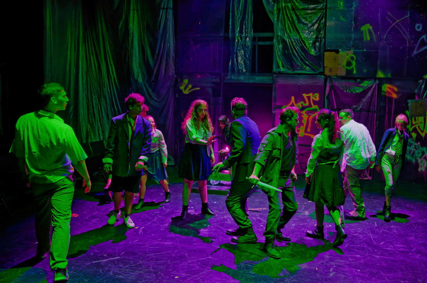 A group of young performers on stage under colorful neon lighting, with graffiti-style artwork in the background, possibly rehearsing or performing.