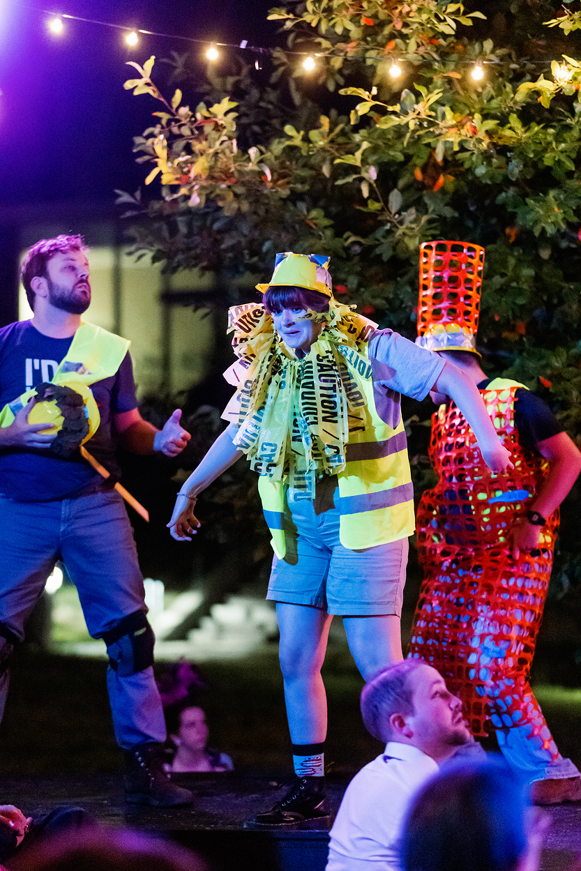 People dressed in colorful, makeshift hazard and caution costumes performing on an outdoor stage at night, with trees and string lights in the background.