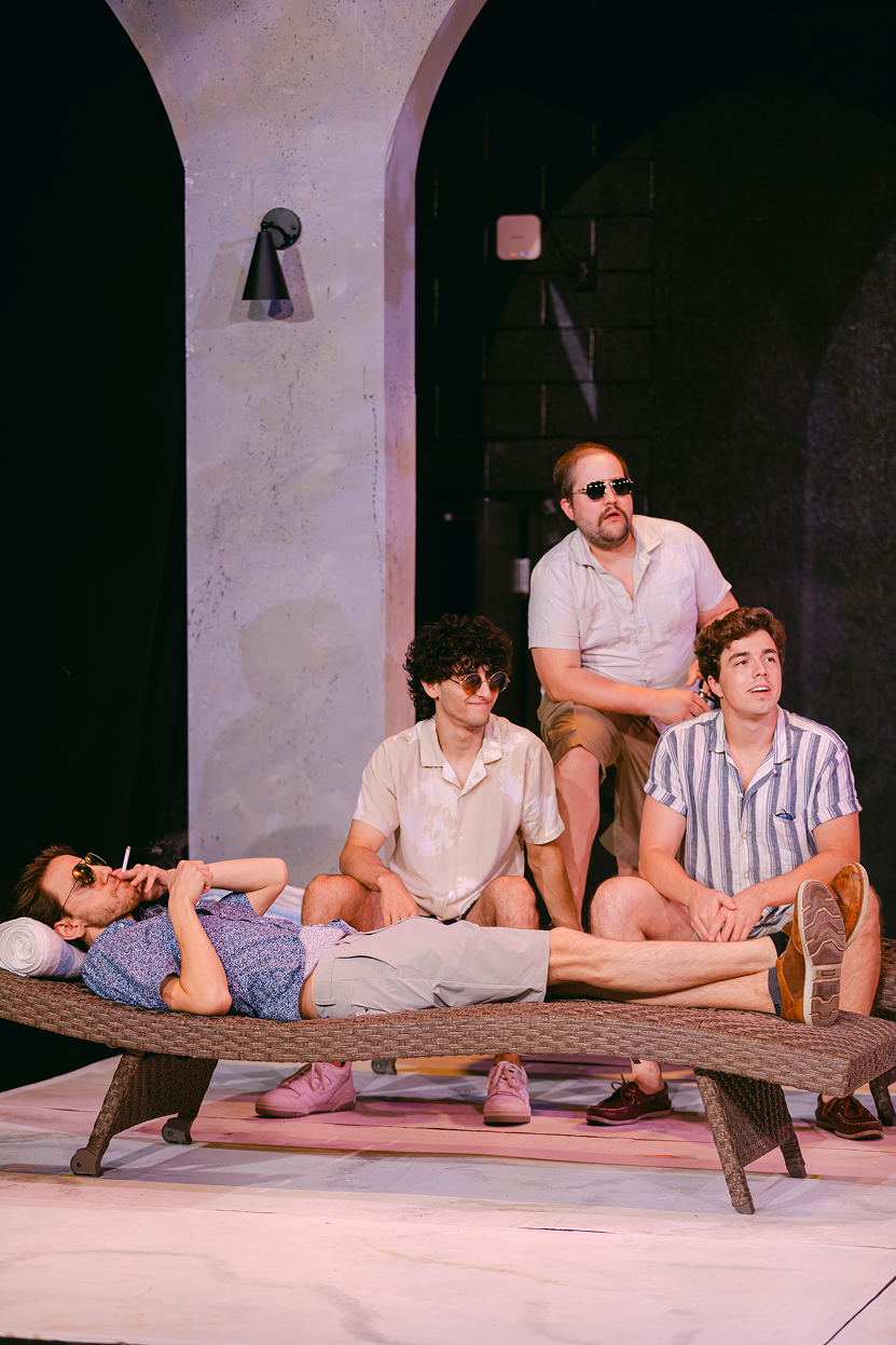 Four men are on stage, with one lying on an outdoor lounge chair and the other three seated or standing around him. They are wearing casual summer clothes and sunglasses. The background features a stone wall and an arched doorway, indicating an indoo