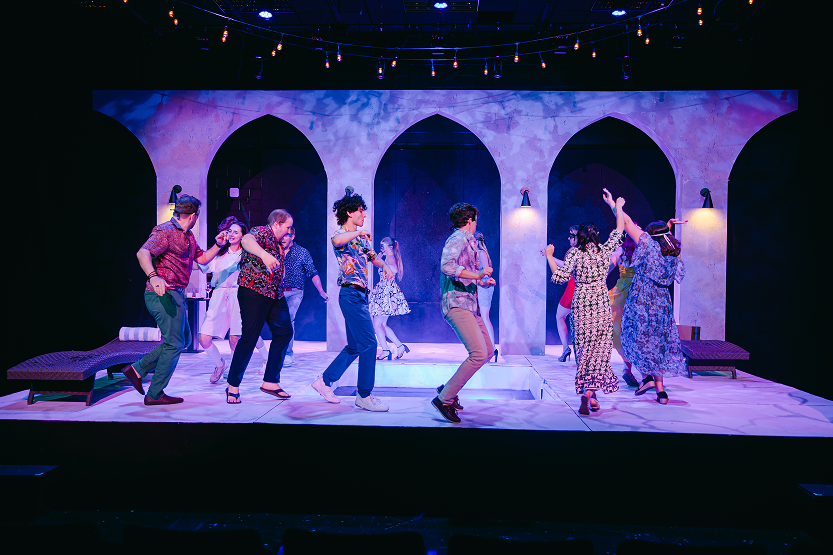 Theater stage with cast dancing during a performance, set to resemble a Mediterranean village with arches, wall sconces, and string lights.