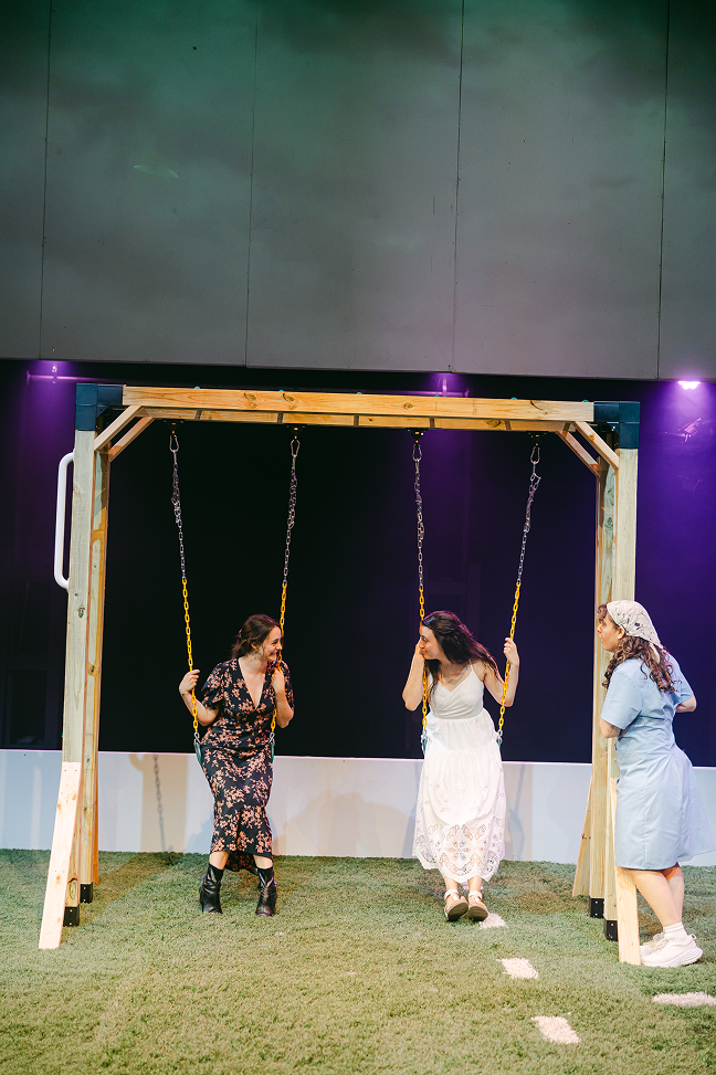 Three women are on a wooden swing set, with one swinging and two standing nearby on artificial grass.