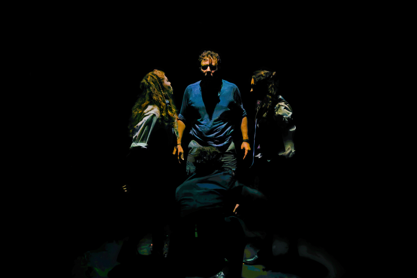Three people standing around a fourth person who is sitting or lying down, in a dark environment with focused lighting on them.