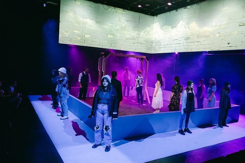 The image shows an indoor theater scene with multiple actors on a stage enclosed by a low wall. Some actors are standing along the wall while others are inside the stage area. The background has a brick wall projection and purple lighting with fog ef