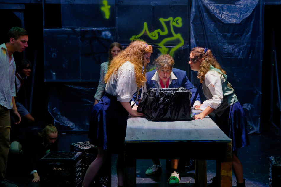 Teen actors performing a scene with a table and black bag on stage, in a theater with dark backdrop and graffiti-style art